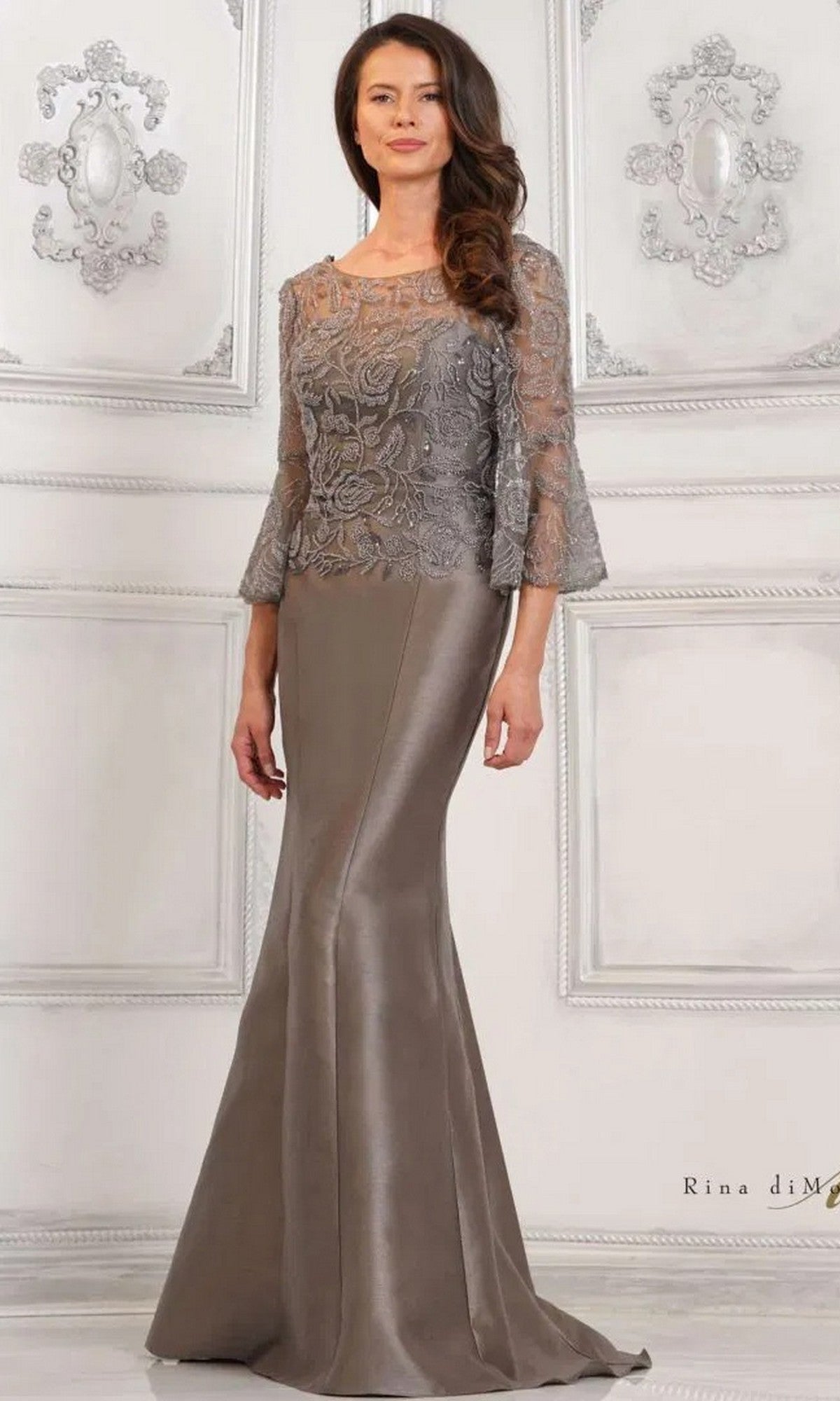 A woman in the gunmetal Rina Di Montella RD2939 long formal dress, with a fitted satin mermaid skirt and lace three-quarter sleeve top, stands against an ornate white wall.